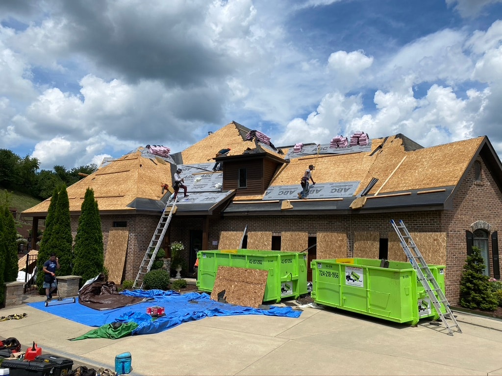 Large brick home during roof tear-off with crew and dumpsters on site